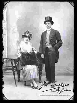 Foto de estudio de una pareja. Una dama que se encuentra sentada en un sillón y que lleva puesto un sombrero con adornos, traje formal con falda, botines con tacón. El varón se encuentra parado y lleva puesto un sombrero tarro, traje frac y zapatos guets.