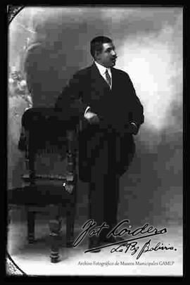 Foto de estudio de un varón de bigote, vestido de traje. Se encuentra parado y apoyado con el brazo derecho a un sillón