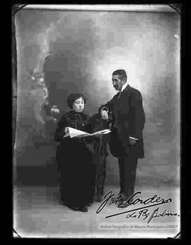 Foto de estudio de una pareja que se encuentran observando una revista. La dama se encuentra sentada en un sillón y lleva puesto un vestido largo de cuello cerrado y de color oscuro. El varón se encuentra parado y lleva puesto un traje formal.