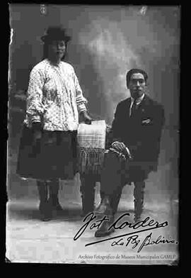 Foto de estudio de una pareja. Una chola que se encuentra parada y apoyada al sillón donde se encuentra sentado su pareja vestido de traje y corbata. Ella lleva puesto un sombrero borsalino de color negro. Una blusa blanca con puntos de cuello cerrado y manga larga, una pollera y botines con tacón.