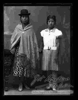 Foto de estudio de una chola que se encuentra parada junto a su hija y apoyada a un sillón. Lleva puesto un sombrero borsalino de color oscuro, manta bordada, pollera con figuras y botines con tacón.