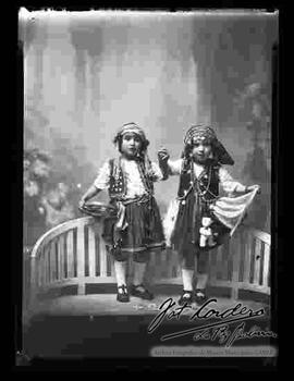 Foto de estudio de dos niñas disfrazadas de gitanas y paradas sobre un sillón de madera tallada.