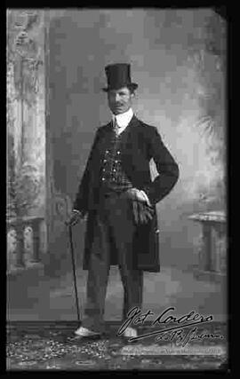 Foto de estudio de un varón que  lleva puesto: Un traje formal con chaleco a cuadros, camisa blanca, corbata, sombrero tarro y zapatos guets. Sostiene en mano un bastón y guantes.