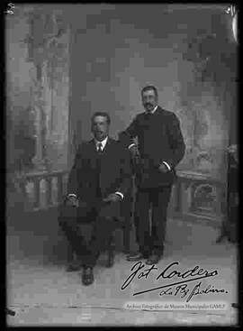 Foto de estudio de dos varones que visten de traje. Uno se encuentra sentado en un sillón y el otro parado a su lado