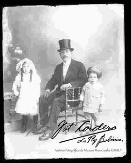 Foto de estudio  de un padre sentado en una silla tallada de madera, vestido de traje y sombrero, junto a sus dos hijos