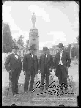 Cuatro varones vestidos de y traje y con sombrero bom bin, delante del monumento a los  Colorados de Bolivia, en el final de la alameda