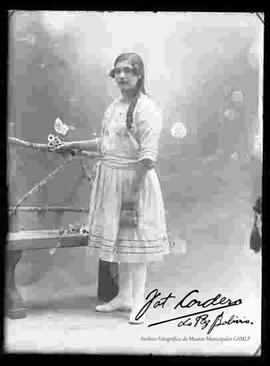 Foto de estudio de una señorita apoyada a una banca y que lleva puesto un vestido blanco, sosteniendo en mano una pequeña cartera.