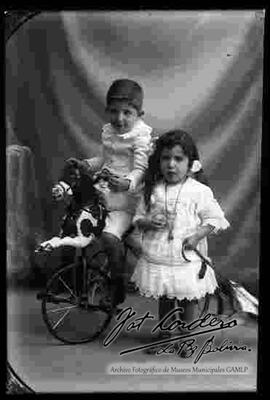 Foto de estudio de dos niños. Una niña y un niño que se encuentra montado en un triciclo de caballito