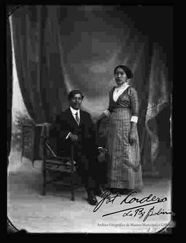 Foto de estudio de una pareja. Un varón que se encuentra sentado en una silla y que lleva puesto un traje formal. La dama se encuentra parada y lleva puesto un vestido largo a rayas.