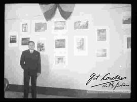 Vista de un varón que viste de traje y que se encuentra posando delante de las fotografías de exposición "Foto Cordero Jiménez", en el salón de la cancillería. octubre 1948.