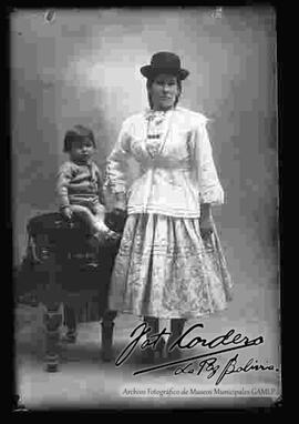 Foto de estudio de una chola que se encuentra parada y apoyada a un sillón donde se encuentra sentado un  niño. Lleva puesto un sombrero borsalino de color negro, una blusa blanca de cuello cerrado manga larga con encajes, una pollera floreada y botines con tacón