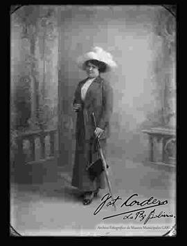 Foto de estudio de una dama que lleva puesto un traje formal, complementado por un sombrero con plumas, una sombrilla, una cartera y botines con tacón.