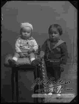 Foto de estudio de dos niños. Uno se encuentra sentado encima de un cojín en una mesita y el otro parado encima de una silla