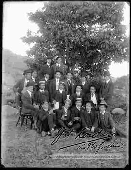 Grupo de varones vestidos de traje y sombrero, compartiendo bebidas en el campo, delante de un árbol