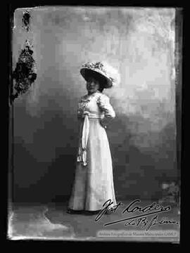 Foto de estudio de una dama que lleva puesto: Un sombrero adornado con flores y pluma, un vestido largo y sujeto con un rosón arriba de la cintura.