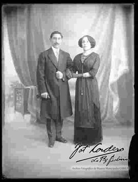 Foto de estudio de una pareja que se encuentran tomados del brazo. Un varón que lleva puesto un traje formal con sacón largo y una dama que lleva puesto un vestido de fiesta largo de color oscuro y  guantes en las manos.