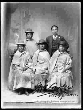 Foto de estudio de cuatro cholas y un varón. Ellas  lucen sombreros borsalinos de color oscuro, mantas puestas, polleras y zapatillas planas. El  varón luce un traje con chaqueta y corbata.