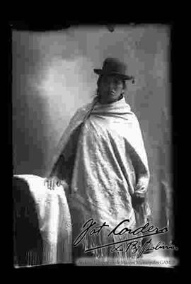 Foto de estudio de una chola que se encuentra parada y apoyada a un sillón donde esta una manta. Lleva puesto un sombrero borsalino de color oscuro, manta bordada y pollera.