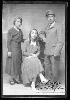 Foto de estudio de tres jóvenes. Dos señoritas que llevan puesto vestidos largos y zapatillas con tacón, un joven que lleva puesto un traje con abrigo y boina en la cabeza.