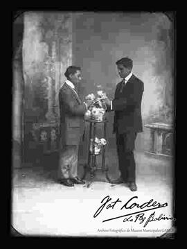 Foto de estudio de dos jóvenes  observando las flores de un florero. Llevan puesto trajes formales.