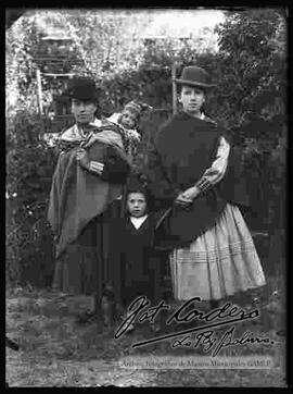 Imagen de dos cholas de provincia junto a sus hijos en el jardín de una casa particular. Llevan puesto sombreros borsalinos de color oscuro, manta, pollera y zapatillas planas