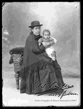 Foto de estudio de una chola que se encuentra sentada en un sillón y sosteniendo a un bebe. Lleva puesto un sombrero borsalino de color oscuro, manta tejida, pollera y botines con tacón