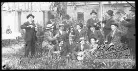 Grupo de varones vestidos de traje, celebrando un acontecimiento, con música y compartiendo bebidas en el huerto de una casa particular