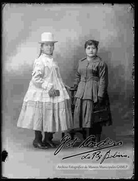 Foto estudio de una chola, que lleva puesto un sombrero borsalino, blusa larga, pollera, anillos en la mano y botines con tacón alto. Sostiene en mano una escarcela (cartera) y junto a ella se encuentra parada una joven de vestido  que porta en mano una cartera