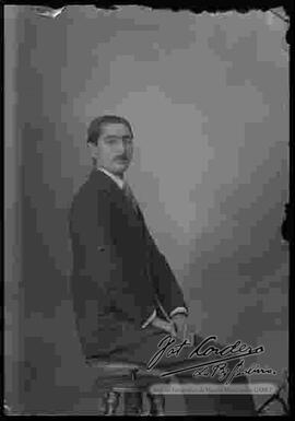 Foto de estudio de un varón que presenta un modelo o tipo de bigote y que se encuentra sentado.