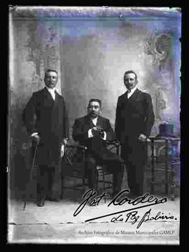 Foto de estudio de tres varones vestidos de  camisa corbata y sacones, sostienen en mano bastones. Dos se encuentran parados y uno sentado en el centro, en una silla artesanal.