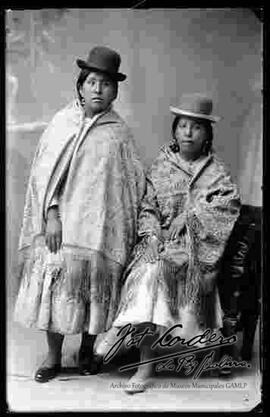 Foto de estudio de dos cholas. Una se encuentra parada y la otra sentada en un sillón. Llevan puestos sombreros  borsalinos de color negro, mantas bordadas, pollera y zapatillas planas