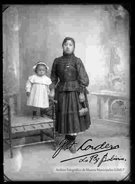 Foto de estudio de una cholita que se encuentra de pie. Lleva puesto una blusa de manga larga con encajes, pollera con enaguas sobresalidas, joyas y con botines de tacón. Sostiene en mano una escarcela (cartera) y junto a ella se encuentra una niña parada sobre un sillón de mimbre.