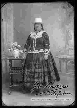 Foto de estudio de una chola que se encuentra parada y apoyada a una mesita con flores. Lleva puesto, un sombrero blanco de copa alta, una blusa cerrada y pollera floreada de tela gamuza, con botines con tacón.