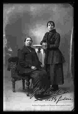 Foto de estudio de dos damas. Una se encuentra parada y la otra sentada en un sillón