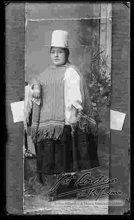 Reproducción de una foto de estudio. Una  chola  que se encuentra parada y apoyada en un mueble. Lleva puesto un sombrero de copa alta color blanco, manta tejida de lana, blusa blanca de manga larga, pollera color negro y botines con tacón