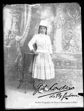 Foto de estudio de una chola que luce, un sombrero copa alta, blusa de cuello cerrado mangas largas pollera con enaguas largas y con botines, apoyada en una mesita