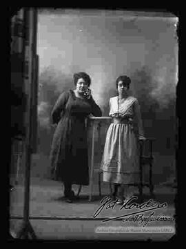 Foto de estudio de dos damas (madre e hija), paradas junto a unos muebles. Llevan puesto vestidos de diferente modelo y color.