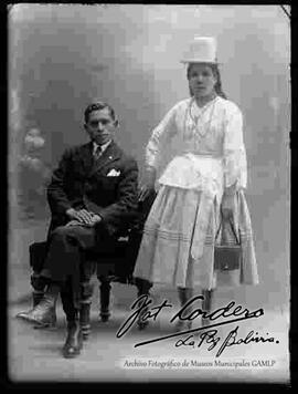 Foto de estudio de una chola junto a su esposo que se encuentra sentado en un sillón. Ella lleva puesto un sombrero blanco copa alta, blusa de manga larga, pollera, botines con tacón, agarrando en la mano izquierda escarcela (cartera)