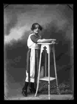 Foto de estudio de una señorita que se encuentra parada y apoyada a una mesita larga. Lleva puesto un vestido de dos colores adornado con seda.