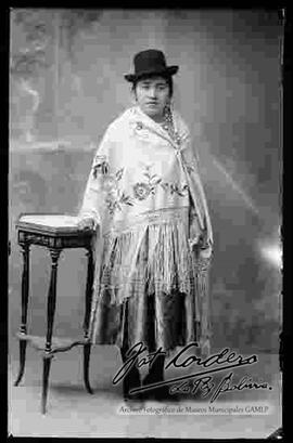 Foto de estudio de una chola que se encuentra parada y apoyada en una mesita. Lleva puesto un  sombrero borsalino de color negro, manta bordada con flores y prendida con topo (prendedor), pollera y botines con tacón