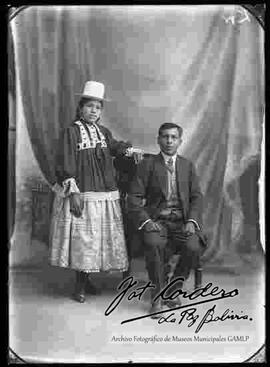 Foto de estudio de una pareja. Una chola que se encuentra parada y que lleva puesto un sombrero blanco de copa alta, una blusa maternal de manga larga, una pollera y botines con tacón. A su lado se encuentra un caballero sentado de traje con corbata.