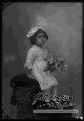 Foto de estudio de una niña que se encuentra sentada en la cabecera o espaldar de un sillón, sosteniendo en mano unas flores