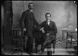 Foto de estudio de dos varones. Uno se encuentra parado y el otro sentado a su lado en un sillón