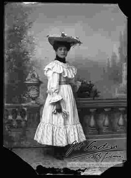 Foto de estudio de una dama que lleva puesto un sombrero adornado con flores,  un vestido largo de color claro y botines con tacón.