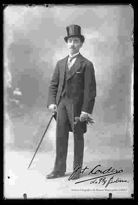 Foto de estudio de un varón que lleva puesto: Un sombrero tarro, camisa blanca, corbata chaleco y un traje frac. Sostiene en las manos unos guantes y un bastón.