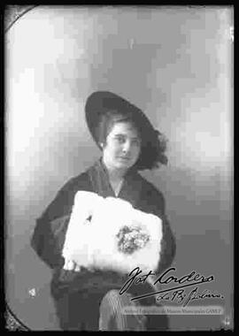 Foto de estudio de una extranjera que se encuentra sentada, sosteniendo una prenda de piel para cubrir las manos. Lleva puesto un sombrero con adornos
