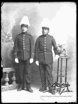 Foto de estudio de dos soldados, parados a lado de un barandal y una mesita con flores