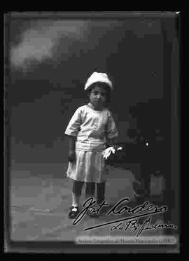 Foto de estudio de un aniña parada junto a un sillón y sosteniendo en la mano izquierda unos guantes blancos