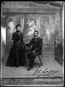 Foto de estudio de una pareja. Una dama que se encuentra parada y que lleva puesto un vestido largo de cuello cerrado y color oscuro. Un varón que se encuentra sentado en un asilla y que viste de traje formal.