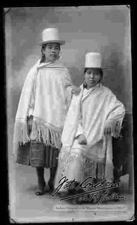 Foto de estudio de dos cholas. Una se encuentra parada y la otra sentada en un sillón. Ambas llevan puesto: Sombreros de copa alta y color blanco, manta, pollera y botines con tacón